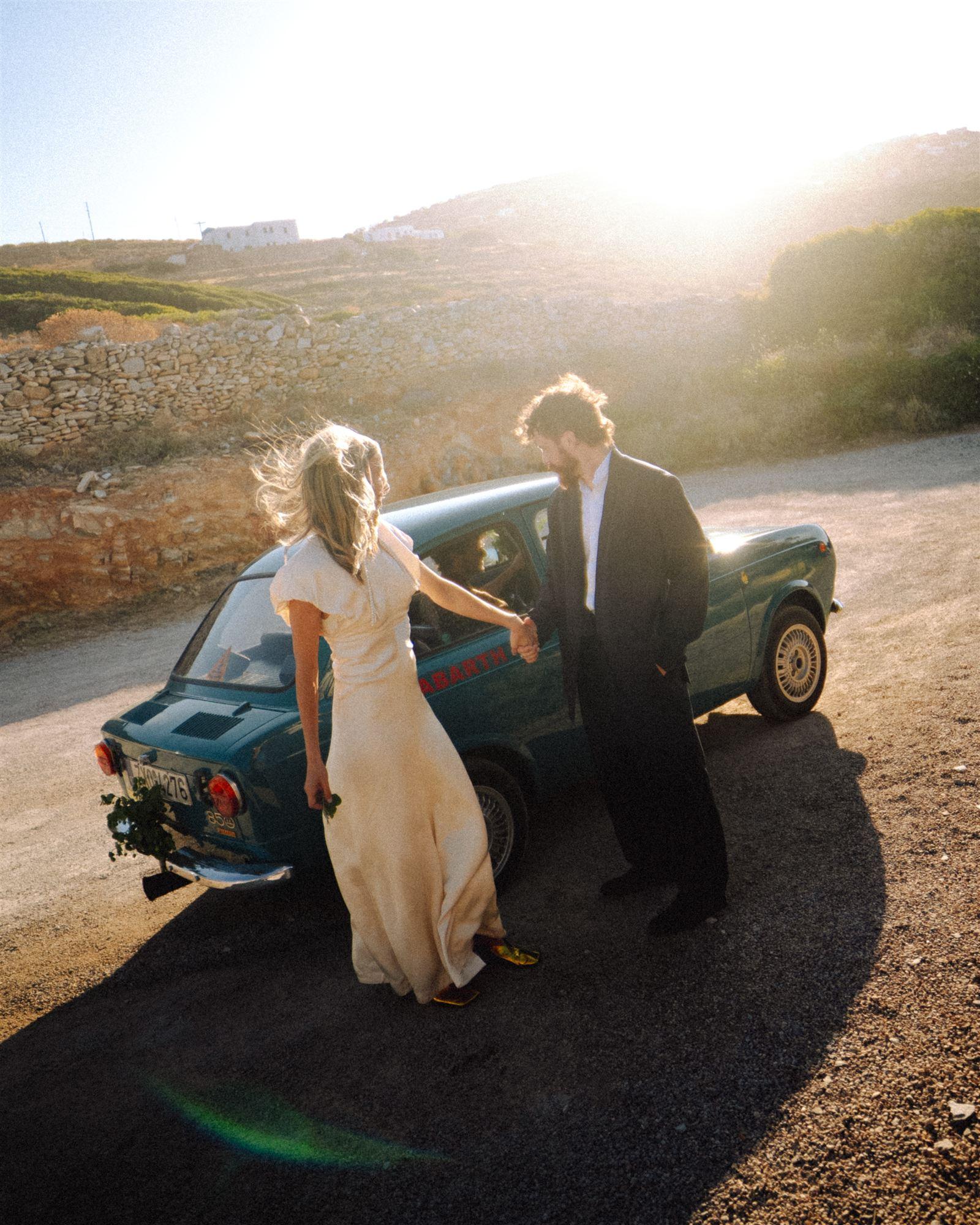 Bride and groom by a vintage car with a family member helping arrange the dress