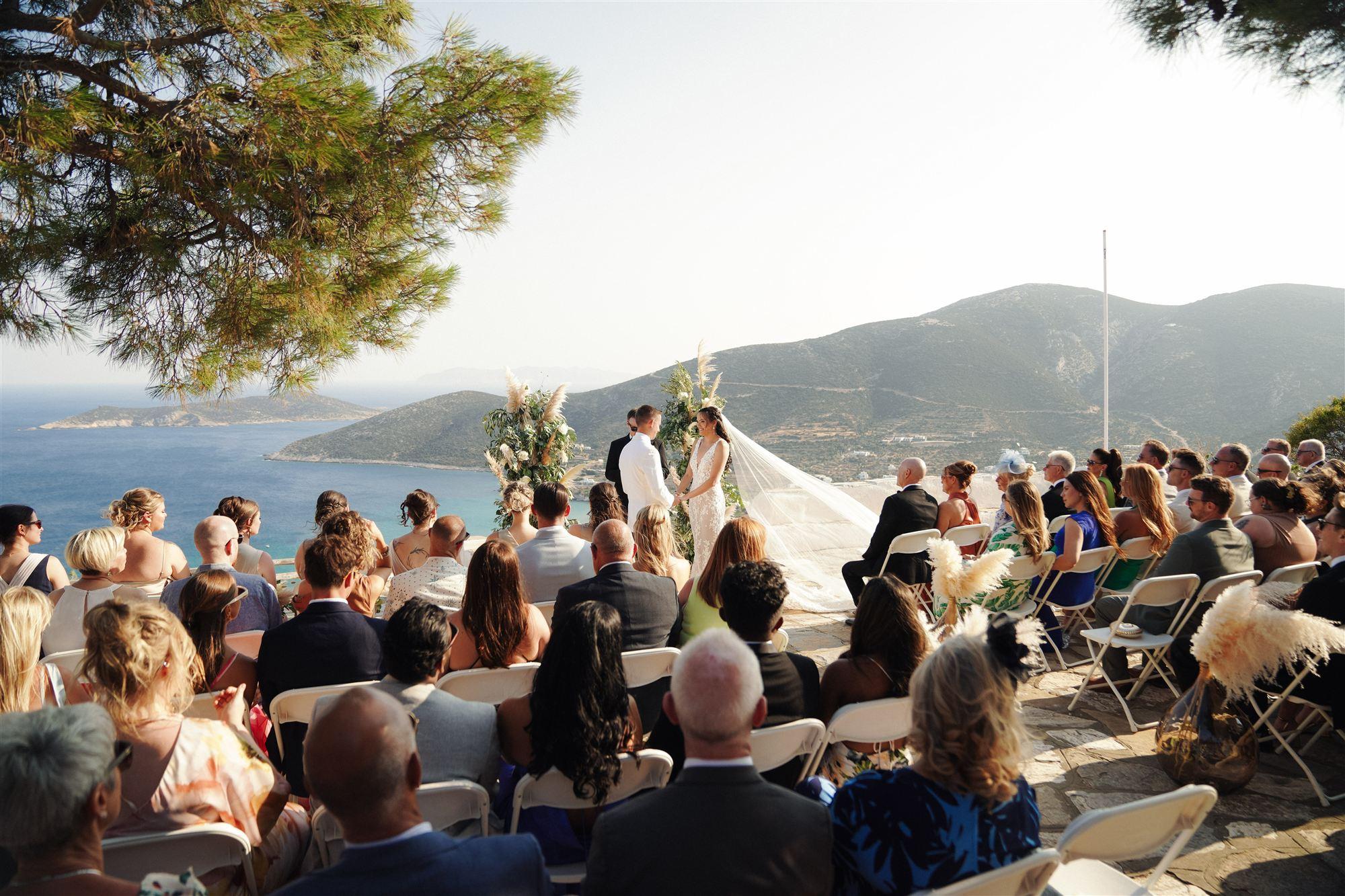 Wedding ceremony on a Sifnos clifftop overlooking the sea, guests seated under pampas grass arrangements
