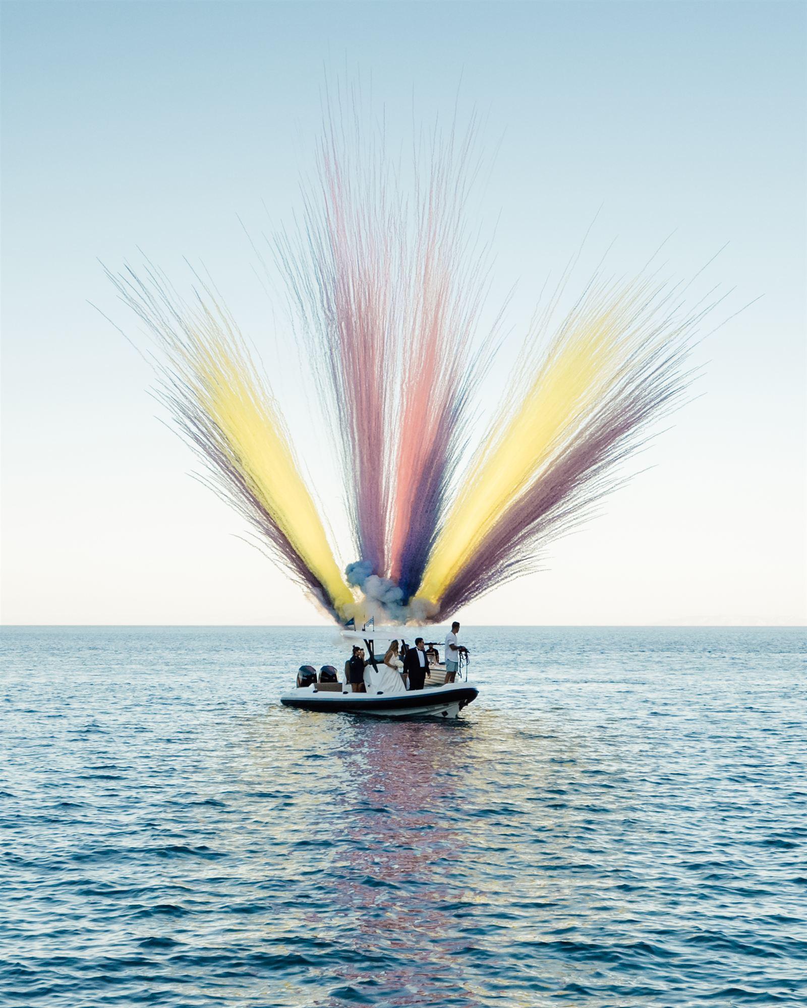 Bride and groom on a boat leaving Sifnos with coloured smoke flares against the sea