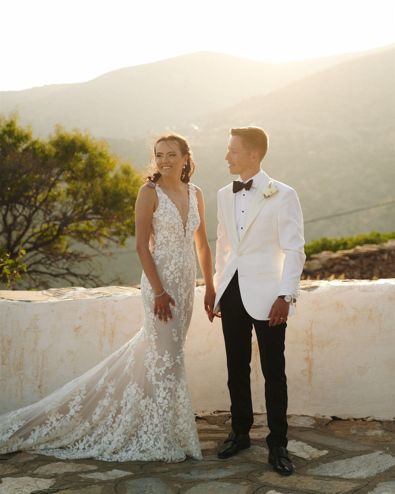 Couple in a white tuxedo dress and jacket, holding hands on a hillside path at golden hour